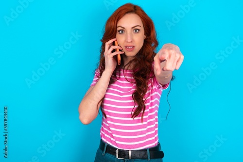 Positive young redhead woman wearing striped T-shirt over blue background indicates directly at camera has telephone conversation smiles broadly enjoys talking long hours. You join me