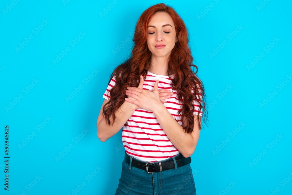 young redhead woman wearing striped T-shirt over blue background smiling with hands on chest with closed eyes and grateful gesture on face. Health concept. young redhead woman wearing striped T-shirt over blue background smiling with hands on chest with closed eyes and grateful gesture on face. Health concept.