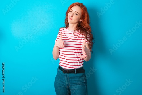 Joyful young redhead woman wearing striped T-shirt over blue background wink and points index fingers at camera, chooses someone, makes finger gun pistol.