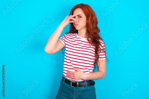 young redhead woman wearing striped T-shirt over blue background smelling something stinky and disgusting, intolerable smell, holding breath with fingers on nose. Bad smell