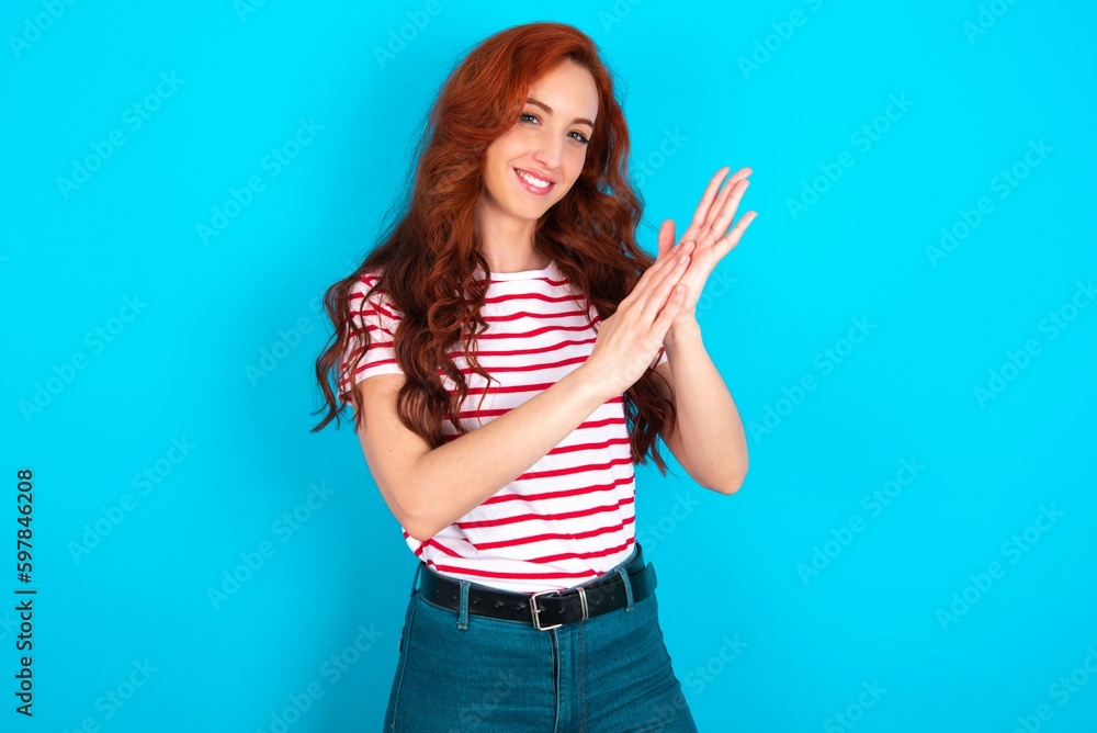 young redhead woman wearing striped T-shirt over blue background clapping and applauding happy and joyful, smiling proud hands together. young redhead woman wearing striped T-shirt over blue background clapping and applauding happy and joyful, smiling proud hands together.