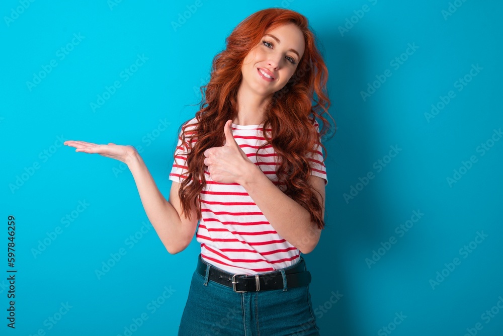 young redhead woman wearing striped T-shirt over blue background Showing palm hand and doing ok gesture with thumbs up, smiling happy and cheerful. young redhead woman wearing striped T-shirt over blue background Showing palm hand and doing ok gesture with thumbs up, smiling happy and cheerful.