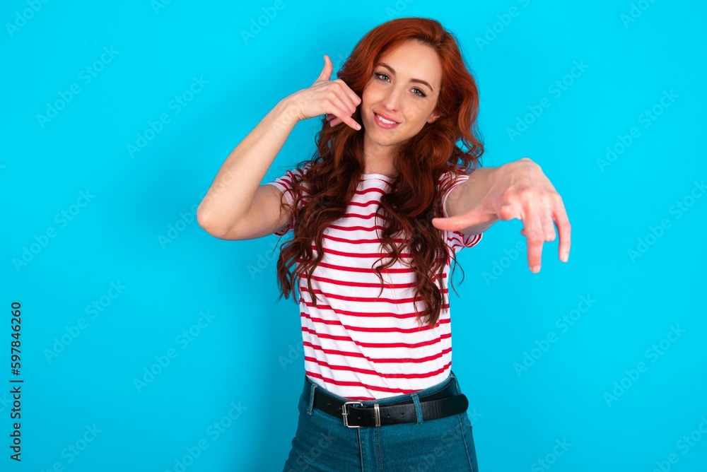 young redhead woman wearing striped T-shirt over blue background smiling cheerfully and pointing to camera while making a call you later gesture, talking on phone young redhead woman wearing striped T-shirt over blue background smiling cheerfully and pointing to camera while making a call you later gesture, talking on phone