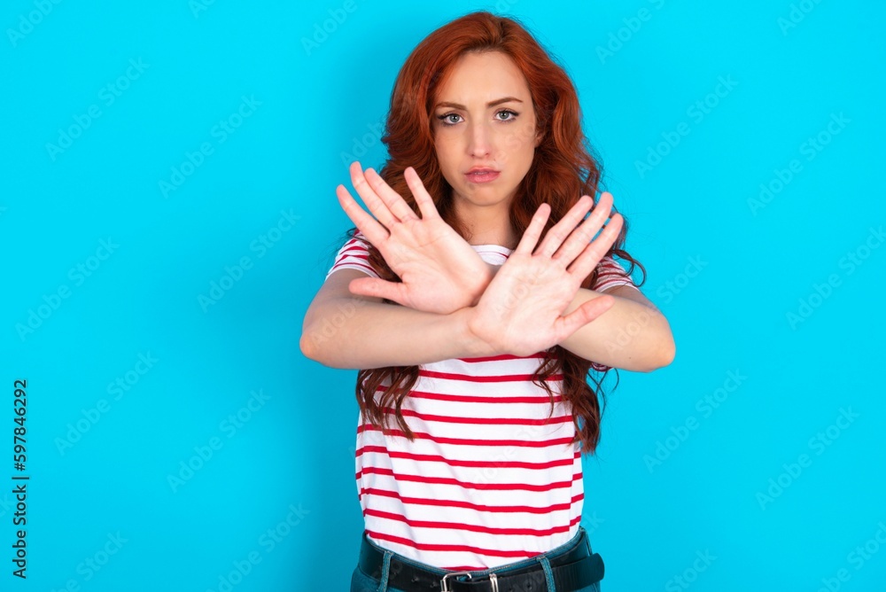 young redhead woman wearing striped T-shirt over blue background has rejection expression crossing arms and palms doing negative sign, angry face. young redhead woman wearing striped T-shirt over blue background has rejection expression crossing arms and palms doing negative sign, angry face.