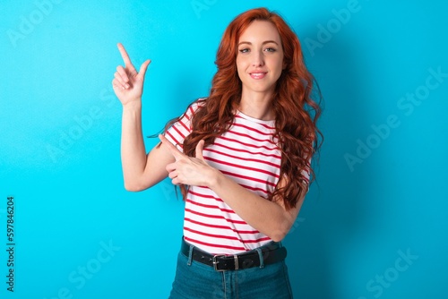 young redhead woman wearing striped T-shirt over blue background indicating finger empty space showing best low prices  looking at the camera