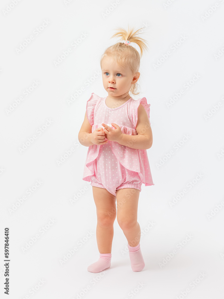 Questioningly serious look. A 1-2-year-old girl in a pink dress and socks stands fingering on a white background. Questioningly serious look. A 1-2-year-old girl in a pink dress and socks stands fingering on a white background.