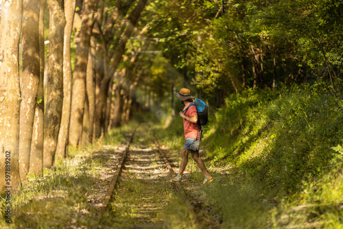 Backpacker man, walking along train tracks, observing Portugal's lush green forest in summertime. Wearing hat, pink t-shirt and short with sandals. Sunbeams.