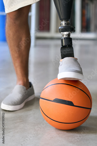 Man amputee with prosthetic leg disability prosthesis standing putting artificial foot on orange basketball ball. Inclusive sport for people with disabilities concept. Vertical close up shot.