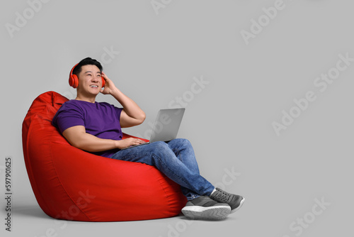 Smiling man with laptop and headphones sitting in beanbag chair against grey background