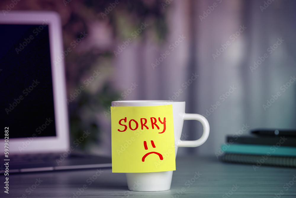 Apology. Sticky note with word Sorry and drawn sad face attached to cup on table in room