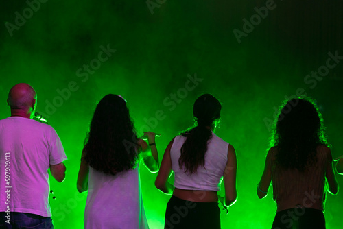 Group of people rehearsing and singing in empty theater seen from the stage, with green light in the background