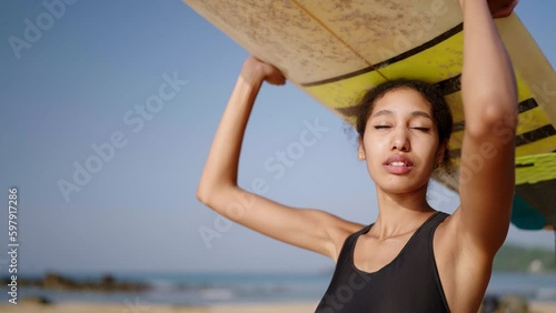 African american surfer standing with surf board on her head on ocean beach. Black woman looking at sea waves on lineup spot. Pretty multiethnic female goes on surfing session, enjoys sun. Slow motion