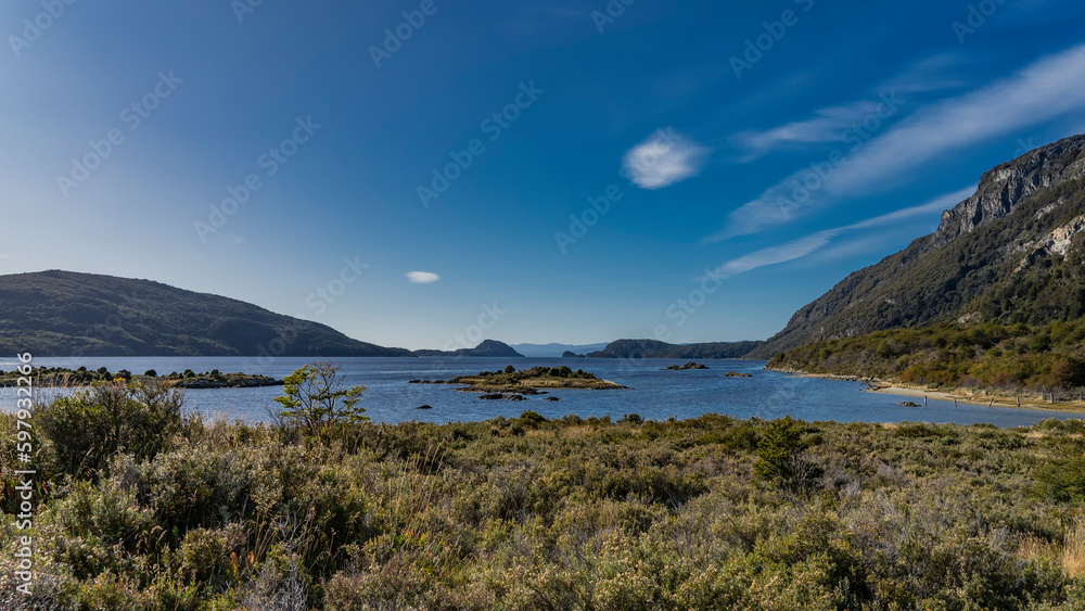 The calm blue lake is surrounded by mountains. Islands in the center. On the shore - low-growing vegetation of Patagonia - grasses, shrubs. Blue sky, clouds. Argentina. Tierra del Fuego