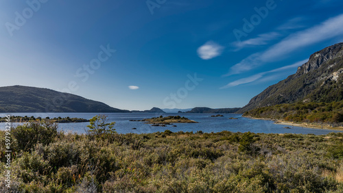 The calm blue lake is surrounded by mountains. Islands in the center. On the shore - low-growing vegetation of Patagonia - grasses, shrubs. Blue sky, clouds. Argentina. Tierra del Fuego