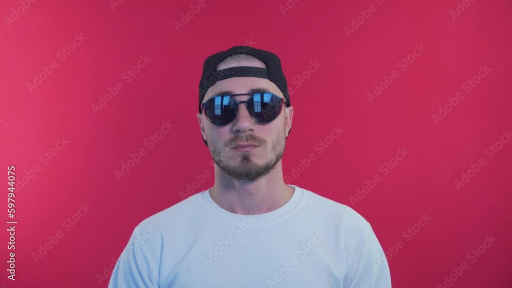 Expressive Young Man in Trendy Sunglasses Smiling and Look Into Camera. Stylish and Fashionable Man on a Red Background in a Baseball Cap and Sunglasses Expressive Young Man in Trendy Sunglasses Smiling and Look Into Camera. Stylish and Fashionable Man on a Red Background in a Baseball Cap and Sunglasses