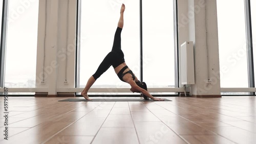 Young caucasian woman in active wear holding Standing Split Pose on yoga mat against background of panoramic window. Urdhva Prasarita Eka Padasana position challenging body focus and balance. photo