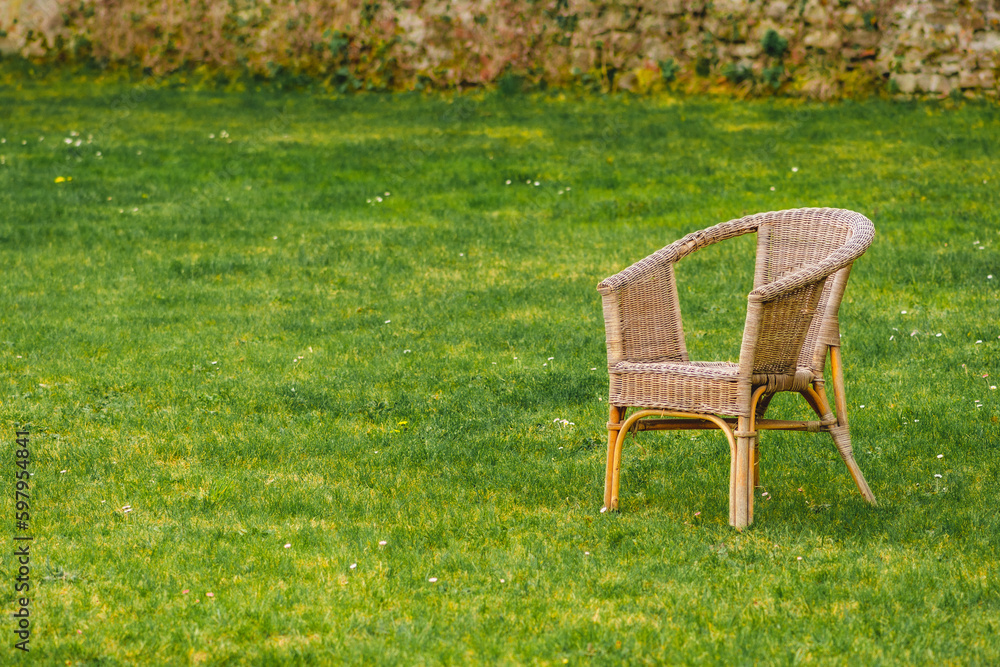 Old empty wicker chair on green grass lawn with flowers and old brick wall on background Old empty wicker chair on green grass lawn with flowers and old brick wall on background