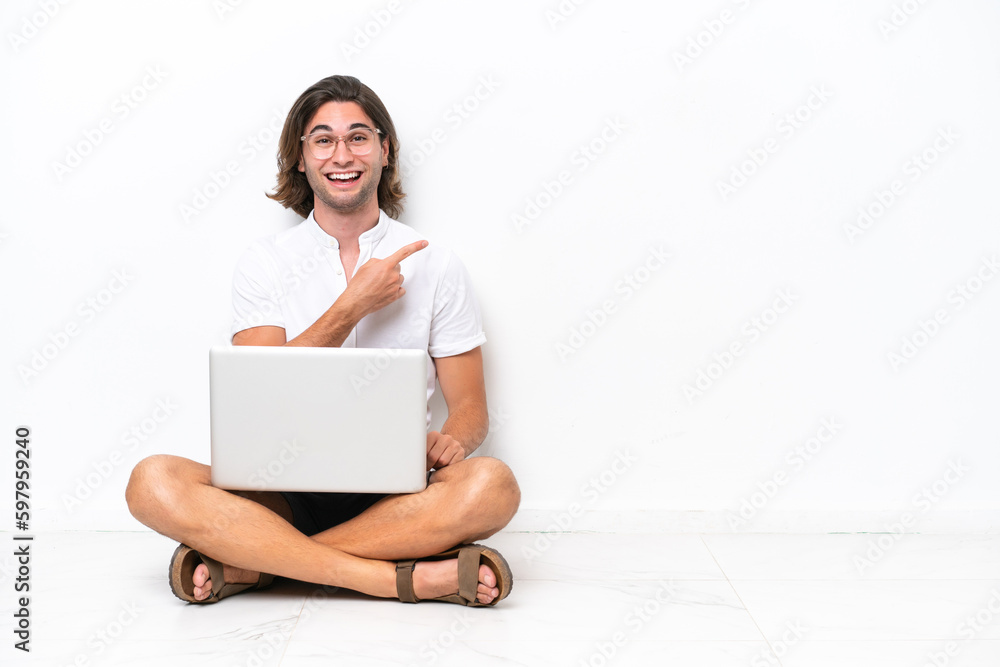 Young handsome man with a laptop sitting on the floor isolated on white background pointing to the side to present a product Young handsome man with a laptop sitting on the floor isolated on white background pointing to the side to present a product