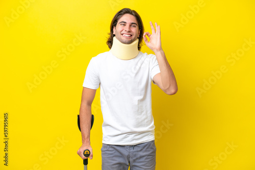 Young handsome man wearing neck brace and crutches isolated on yellow background showing ok sign with fingers