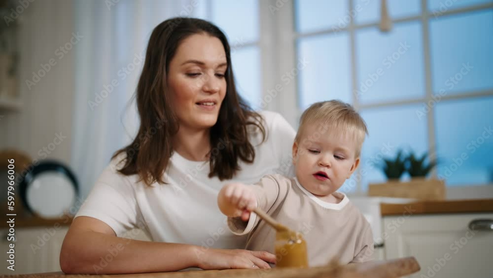 Happy smiling positive mom looks on baby girl eats herself fruit or vegetable puree from jar with spoon sits on kitchen at home. Babyhood, childhood, maternity leave, rising child motherhood concept. Happy smiling positive mom looks on baby girl eats herself fruit or vegetable puree from jar with spoon sits on kitchen at home. Babyhood, childhood, maternity leave, rising child motherhood concept.