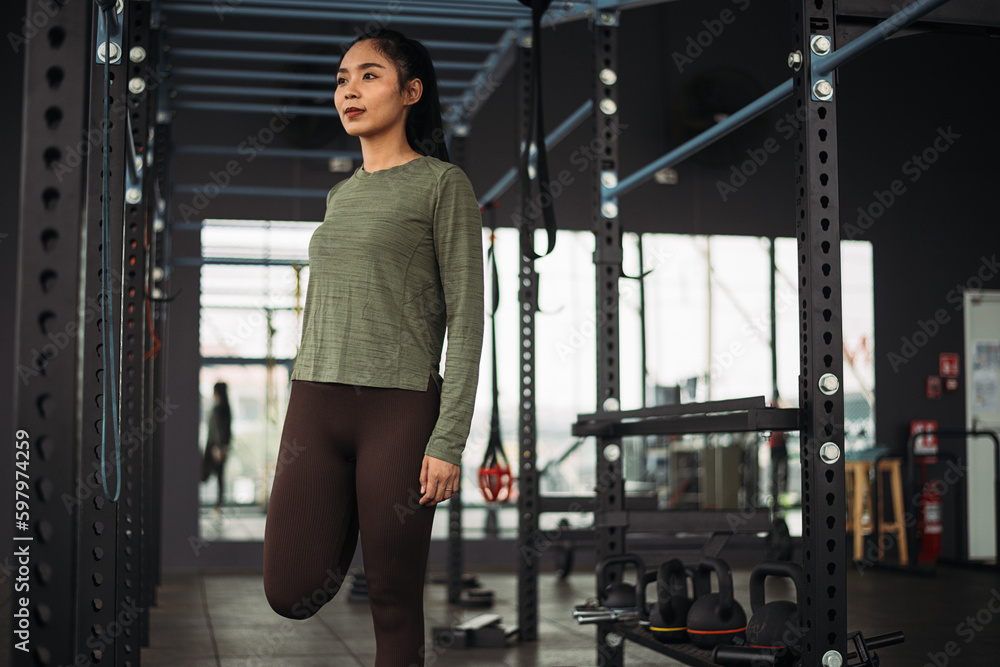 Strong asian woman streching body before exercise at crossfit gym. Athlete female wearing sportswear warm-up before workout on grey gym background with weight and dumbbell equipment. Strong asian woman streching body before exercise at crossfit gym. Athlete female wearing sportswear warm-up before workout on grey gym background with weight and dumbbell equipment.