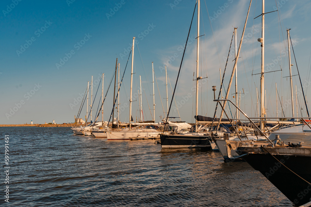 Several boats with masts are docked in the port, on a hot summer evening. In the background you can see the Genoese lighthouse and an old industrial platform