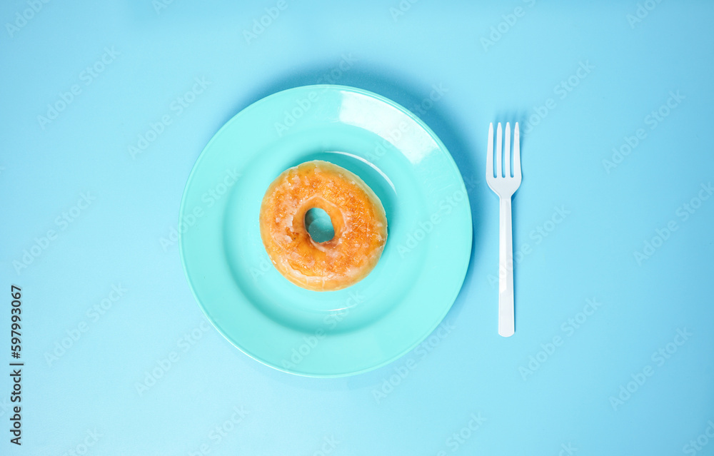 Donut on blue plate with white plastic fork on blue background, top view junk food concept.
