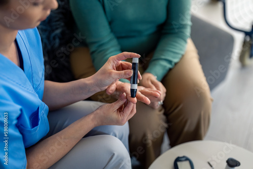 Unrecognizable female nurse poking patient's finger with needle pen to measure blood sugar during home visit. Caregiver using lancet on finger to check patient's blood sugar level by glucose meter. photo