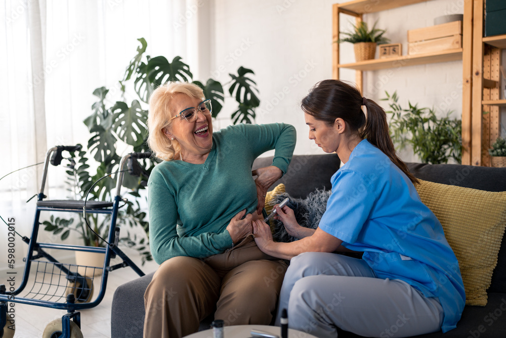 Obraz premium Young dedicated nurse giving insulin injection to a diabetic female patient during home visit. Satisfied smiling senior woman receiving her daily therapy for blood sugar treatment at home.