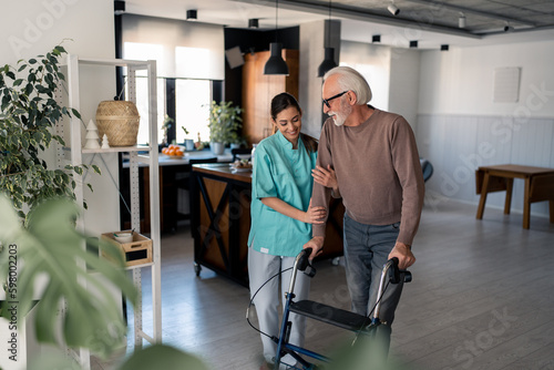 Caring physiotherapist assisting elderly male patient with glasses to walk step by step with mobility walker during home visit. Supportive female physiotherapist helping old man to use mobility walker