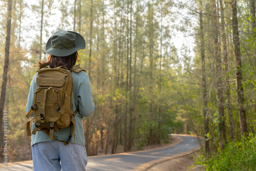 Young Asian girl with a backpack and hat hiking in the mountains during the summer season, a traveler walking in the forest. Travel, adventure, and journey concept.