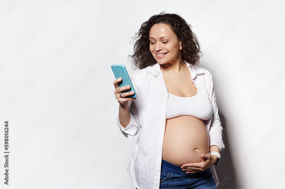 Curly haired multi ethnic brunette, cheerful happy woman in 30 weeks of pregnancy smiling while recording baby kicks on mobile application in her smartphone, isolated white background. Copy ad space Curly haired multi ethnic brunette, cheerful happy woman in 30 weeks of pregnancy smiling while recording baby kicks on mobile application in her smartphone, isolated white background. Copy ad space