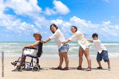 Happy disabled senior elderly woman in wheelchair spending time together with family on holiday beach. Asian grandma, daughter, grandchild boy standing in line, put hands on waists of person ahead.