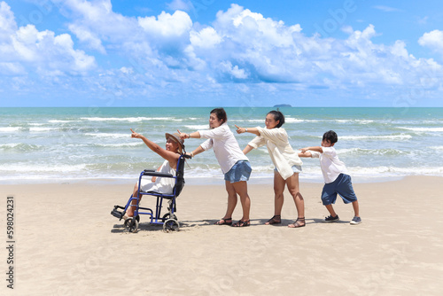 Happy disabled senior elderly woman in wheelchair spending time together with family on holiday beach. Asian grandma, daughter, grandchild boy standing in line, pointing forward, relaxing on holiday.