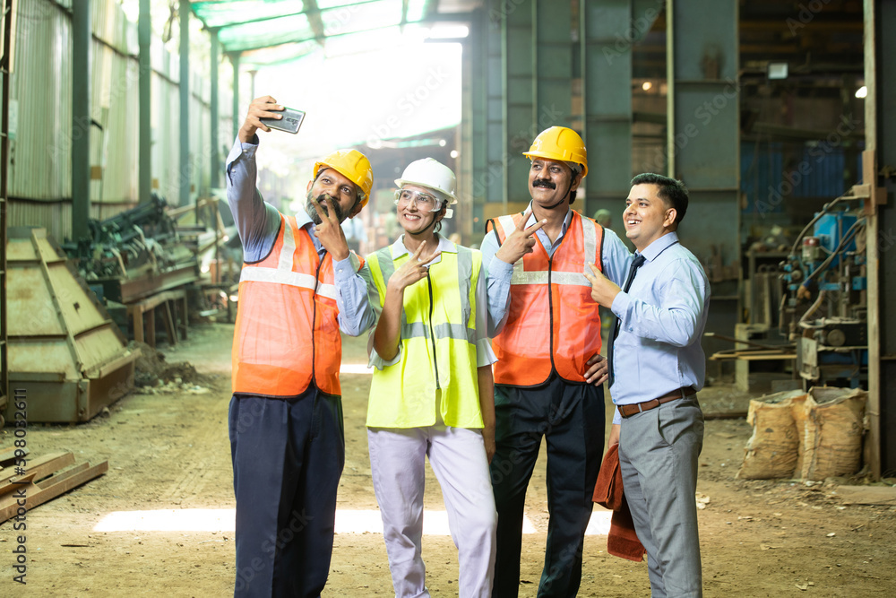 Group of happy indian engineers wearing safety hard hat and vest standing at industrial factory taking selfie picture with smart phone. Work culture. Group of happy indian engineers wearing safety hard hat and vest standing at industrial factory taking selfie picture with smart phone. Work culture.