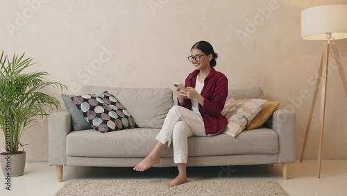 Smiling young woman sitting on a couch at the living room, using mobile phone Generative AI