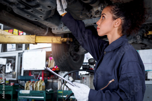 Beautiful female auto mechanic checking wheel tires in garage, car service technician woman repairing customer car at automobile service, inspecting vehicle underbody and suspension engine system. © Stella
