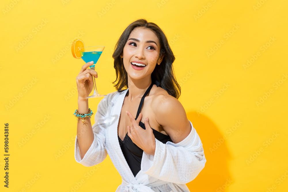 Smiling pretty Asian woman wearing summer swimsuit and spa bathrobe holding cocktail drink in yellow color isolated background studio shot - obrazy, fototapety, plakaty 