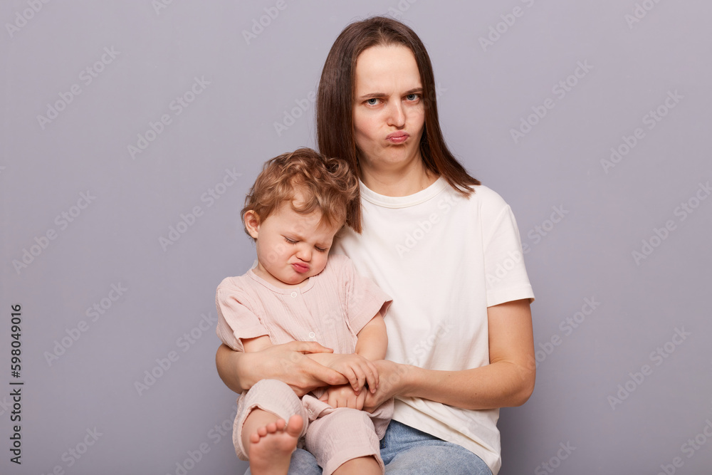Funny confused mother and her infant daughter sitting together isolated over gray background pout lips wearing casual clothing free space for promotion. Funny confused mother and her infant daughter sitting together isolated over gray background pout lips wearing casual clothing free space for promotion.