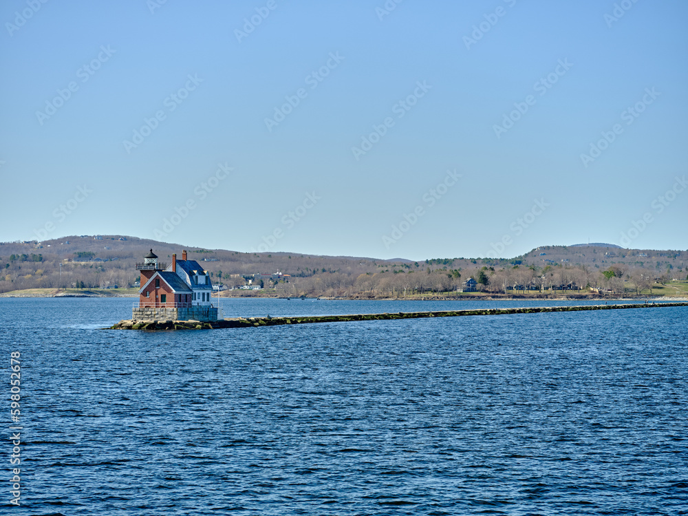 The Rockland Breakwater Lighthouse as seen from the ferry heading back into Rockland harbor with the town of Rockland maine in the background with the rocky breakwater leading back to the shoreline
