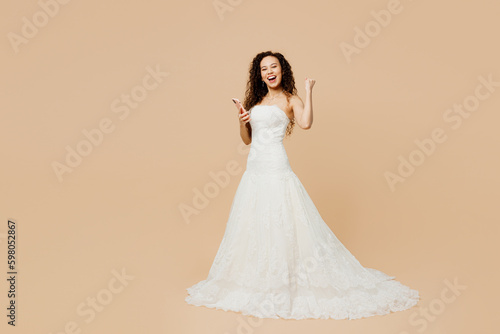 Full body happy young woman bride wear wedding dress posing hold use mobile cell phone do winner gesture isolated on plain pastel beige background studio portrait. Ceremony celebration party concept.