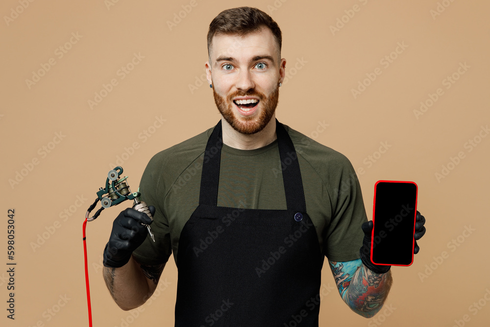 Φωτογραφία Tattooer master tattooed man wear green t-shirt apron use blank screen area mobile cell phone hold machine black ink in jar, equipment for making tattoo art on body isolated on plain beige background