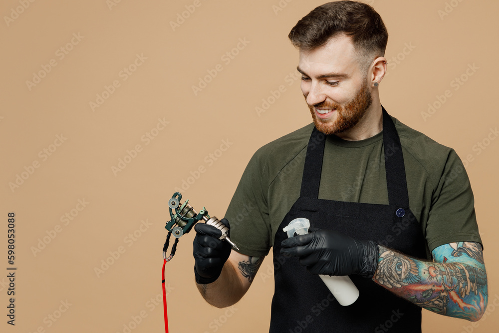 Φωτογραφία Tattooer master artist tattooed man wears green t-shirt apron hold apply antiseptic spray on hand machine black ink in jar, equipment for making tattoo art on body isolated on plain beige background