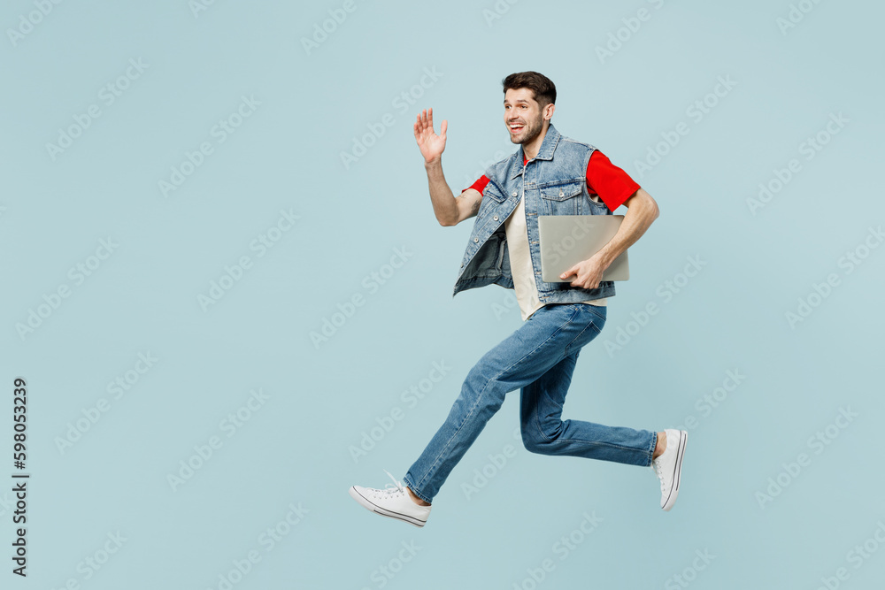 Φωτογραφία Full body side view young IT man wear denim vest red t-shirt casual clothes jump high hold closed laptop pc computer isolated on plain pastel light blue background studio portrait