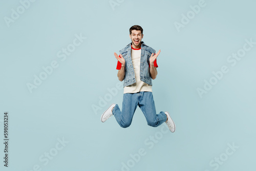 Full body excited happy cool young man he wears denim vest red t-shirt casual clothes jump high spread hands look camera isolated on plain pastel light blue cyan background studio. Lifestyle concept.
