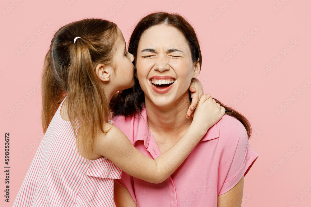 Happy fun adorable lovely woman wearing casual clothes with child kid girl 6-7 years old. Daughter kissing mother cheek, close eyes isolated on plain pastel pink background. Family parent day concept.