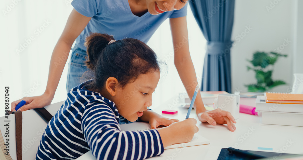Obraz premium Asian toddler girl with sweater sit in front of desk with notepad use pencil focus on write notebook do math homework with her mother or tutor on weekend at home. Distance online learning concept.