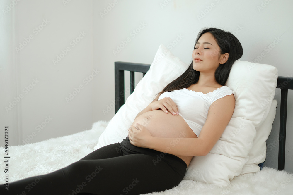 An asian relaxing pregnant woman laying on white bed closing eye with little smile, her hand touching on belly with love emotion. Mother taking a nap on bed. An asian relaxing pregnant woman laying on white bed closing eye with little smile, her hand touching on belly with love emotion. Mother taking a nap on bed.