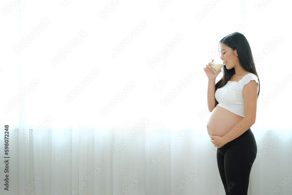 An asian pregnant woman standing and drinking milk at the window with white background and copy space. Her left hand touching at stomarch with love. An asian pregnant woman standing and drinking milk at the window with white background and copy space. Her left hand touching at stomarch with love.