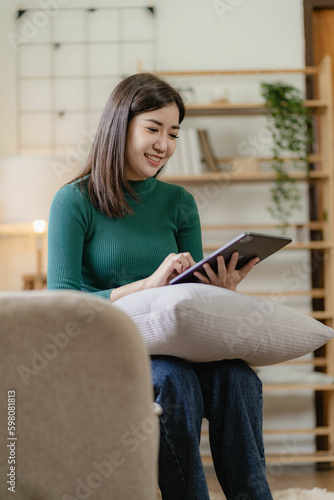Happy asian girl holding tablet device using digital tablet technology sitting on sofa at home smiling woman using app online shopping